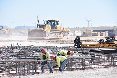 Image of construction workers moving and adjusting rebar on job site with large equipment in the background, moving across a wide open construction site.