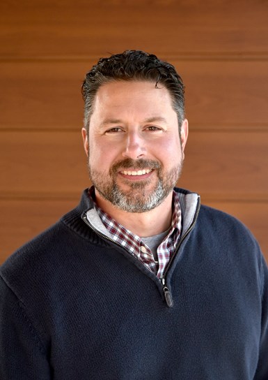 Photo of Chris Jones standing in front of wooden wall wearing dark blue 1/4 zippered sweater with checkered shirt and gray undershirt.